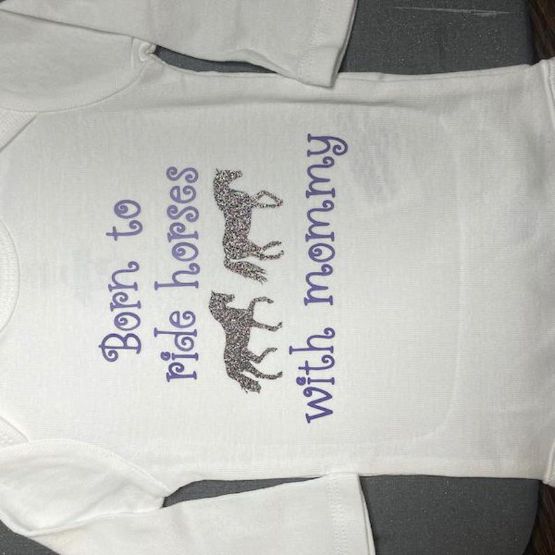 May include: A white long-sleeved baby bodysuit featuring the purple text Born to ride horses with mommy and two glittery silver horse silhouettes positioned between the words ride and horses and the word with.