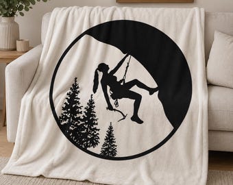 May include: Cream-colored throw blanket featuring a black silhouette of a rock climber within a circular frame. The design includes a climber, rock face, and pine trees. The blanket is draped over a beige sofa.