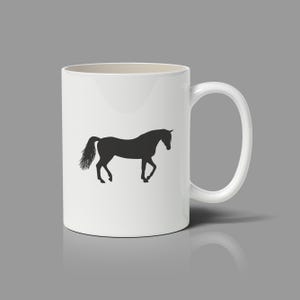May include: A white ceramic mug featuring a black silhouette of a horse standing in profile. The horse is depicted with its head slightly lowered and its tail flowing behind it. The mug has a smooth, cylindrical body and a curved handle on the right.