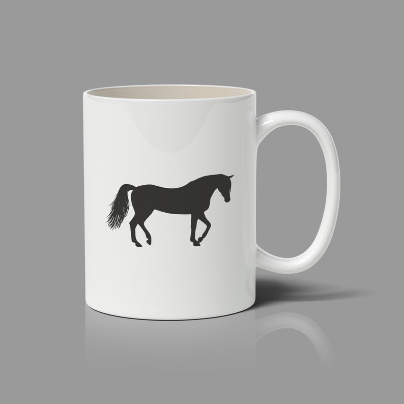 May include: A white ceramic mug featuring a black silhouette of a horse standing in profile. The horse is depicted with its head slightly lowered and its tail flowing behind it. The mug has a smooth, cylindrical body and a curved handle on the right.