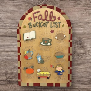 May include: Wooden fall bucket list sign with a red and tan checkered border. The sign features fall-themed illustrations and phrases like "Cozy Book," "Pumpkin Spiced Beverage," and "Visit a Fall Festival."