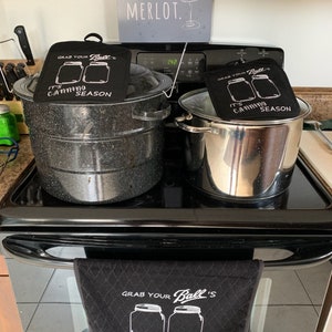 May include: A black stove with two canning pots and a black towel. The towel and two pot holders have the text "GRAB YOUR Ball's IT'S CANNING SEASON" and a graphic of two jars. A sign above the stove says "YOU HAD ME AT MERLOT."