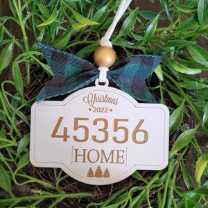 May include: White wooden Christmas ornament with a gold bow, the number 4536, the word "HOME" and a row of three pine trees. The ornament has a white string for hanging and the text "Christmas 2022" at the top.