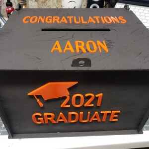 K&ouml;nnte beinhalten: Schwarze Holzkiste mit einer orangefarbenen Abschluss-Kappe, dem Jahr 2021 und dem Wort "Absolvent". Die Kiste tr&auml;gt auch die Worte "Herzlichen Gl&uuml;ckwunsch Aaron" auf der Oberseite.
