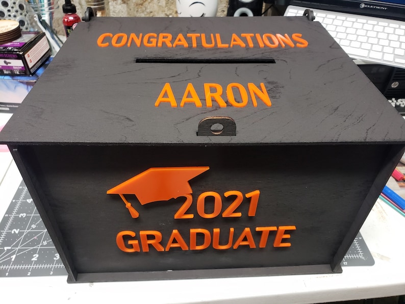 K&ouml;nnte beinhalten: Schwarze Holzkiste mit einer orangefarbenen Abschluss-Kappe, dem Jahr 2021 und dem Wort "Absolvent". Die Kiste tr&auml;gt auch die Worte "Herzlichen Gl&uuml;ckwunsch Aaron" auf der Oberseite.