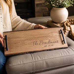 May include: A rectangular wooden serving tray with natural live edges and two matte black metal handles. The surface is engraved with the text The Brockmans and est. 2018 in a script and serif font combination, featuring a light brown wood grain finish.