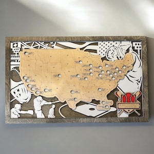 May include: A rectangular wooden wall art piece featuring a layered map of the United States with cutouts for baseball stadiums. The design includes white silhouettes of baseball players, a glove, and a bat emblem with the text THE RYAN'S MLB ADVENTURES CODY & TIA.