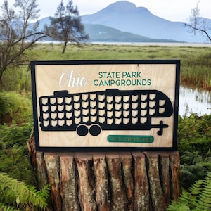 May include: A wooden sign with a black frame, depicting a camper trailer with the state of Ohio outlined on each side. The sign reads "Ohio State Park Campgrounds" and "The Buckinghams".