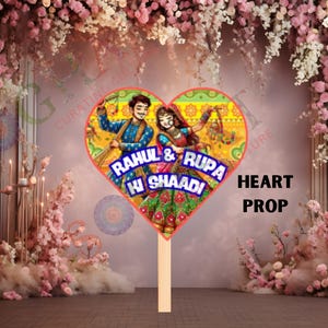 May include: Heart-shaped prop with a colorful illustration of a couple in traditional Indian attire, with the text "RAHUL & RUPA KI SHAADI". The prop is on a wooden stick, with the words "HEART PROP" in black.