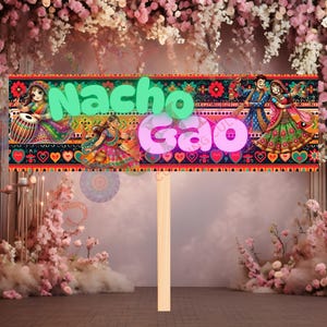 May include: A decorative sign with the words "Nacho Gao" in green and pink, featuring colorful illustrations of Indian dancers and musicians. The sign is mounted on a wooden stick, suitable for celebrations.