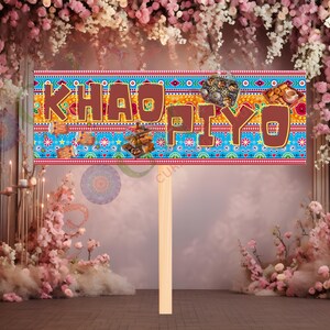 May include: A decorative sign with the words "KHAO PIYO" in brown letters. The sign has a blue background with colorful patterns and images of food. It is held up by a wooden stick, perfect for a party or event.