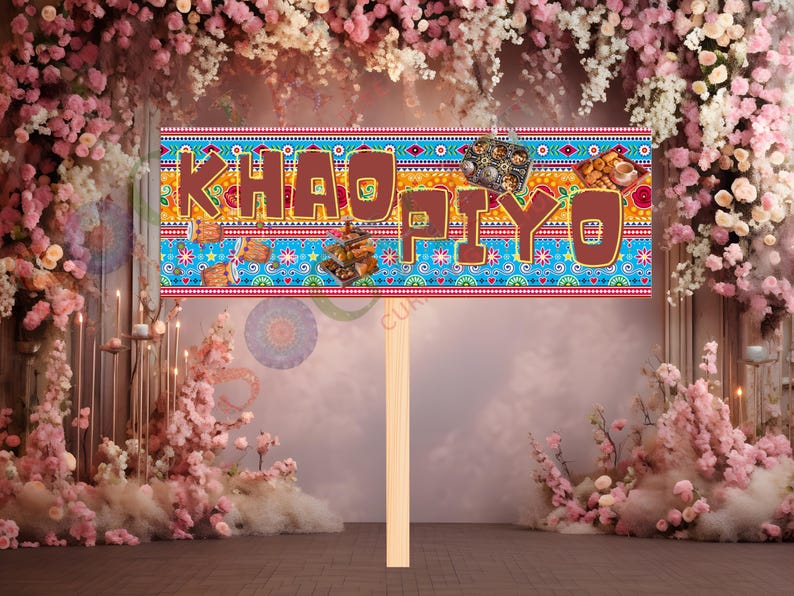 May include: A decorative sign with the words "KHAO PIYO" in brown letters. The sign has a blue background with colorful patterns and images of food. It is held up by a wooden stick, perfect for a party or event.