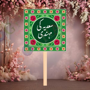 May include: A green square sign with floral and geometric patterns, featuring the text "سعیدہ کی مہندی" in white script. The sign is mounted on a wooden stick, suitable for a wedding or celebration.