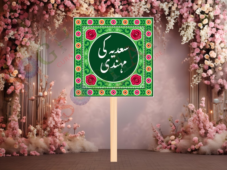 May include: A green square sign with floral and geometric patterns, featuring the text "سعیدہ کی مہندی" in white script. The sign is mounted on a wooden stick, suitable for a wedding or celebration.