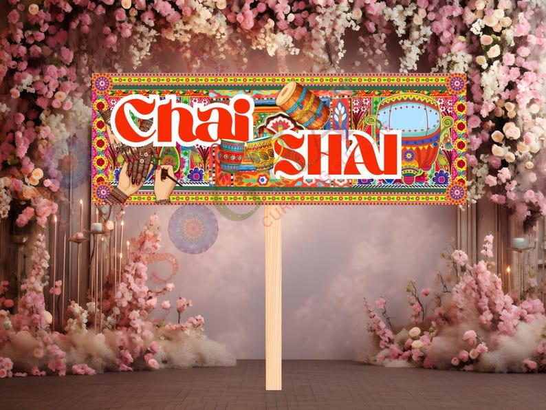 May include: A colorful sign with the words "Chai SHAI" in red, featuring intricate patterns and illustrations of drums and other cultural elements. The sign is held by a wooden stick, set against a backdrop of pink flowers.