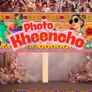 May include: A decorative sign with the text "Photo Kheecho" in red, featuring illustrations of a scooter, a woman with sunglasses, and a photographer. The sign has a colorful, patterned border and is mounted on a wooden post.