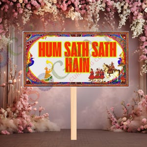 May include: A rectangular sign with the text "HUM SATH SATH HAIN" in orange, framed by a colorful border with traditional Indian motifs. The sign is held by a wooden stick, set against a backdrop of pink and white flowers.