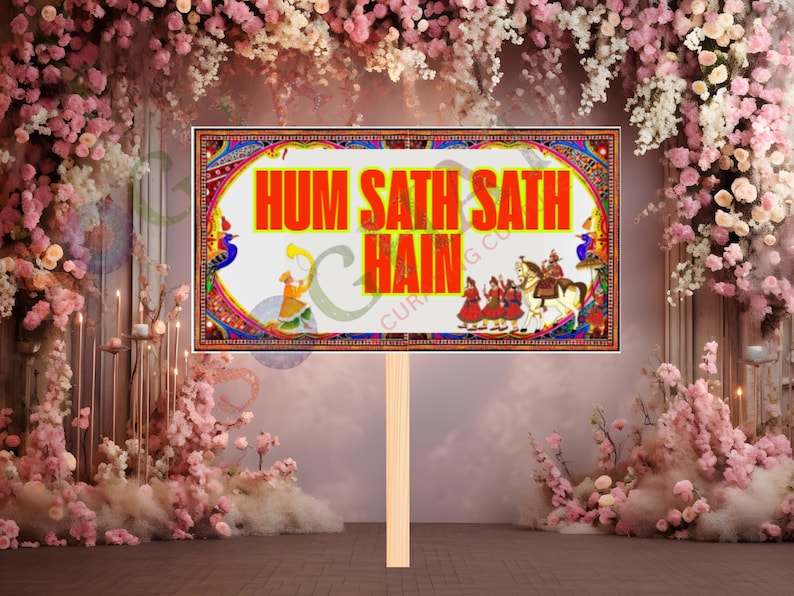 May include: A rectangular sign with the text "HUM SATH SATH HAIN" in orange, framed by a colorful border with traditional Indian motifs. The sign is held by a wooden stick, set against a backdrop of pink and white flowers.
