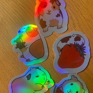 K&ouml;nnte beinhalten: F&uuml;nf schimmernde holografische Aufkleber mit Cartoon-Meerschweinchen mit verschiedenen Gesichtsausdr&uuml;cken und verschiedenen Fr&uuml;chten in den H&auml;nden. Die Meerschweinchen sind braun und wei&szlig; mit schwarzen Augen und Nasen. Die Fr&uuml;chte umfassen eine Erdbeere, eine Wassermelone und einen Kaktus.
