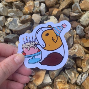 K&ouml;nnte beinhalten: Ein Aufkleber mit einem Cartoon-Meerschweinchen, das einen rosa-blauen Partyhut tr&auml;gt und einen rosa-wei&szlig;en Geburtstagskuchen mit Kerzen h&auml;lt.