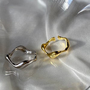 Puede incluir: Dos anillos de forma abstracta, uno plateado y otro dorado, sobre un fondo de tela blanca.