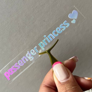 May include: A clear sticker with the words "passenger princess" in iridescent lettering. Two small iridescent hearts are on the right side of the sticker. A gold-colored tool is holding the sticker.