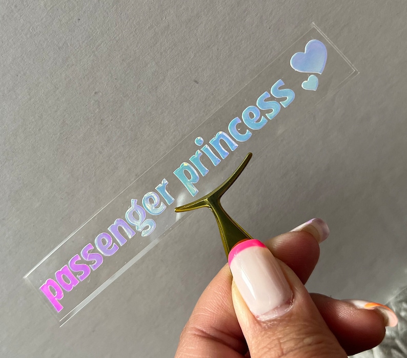 May include: A clear sticker with the words "passenger princess" in iridescent lettering. Two small iridescent hearts are on the right side of the sticker. A gold-colored tool is holding the sticker.