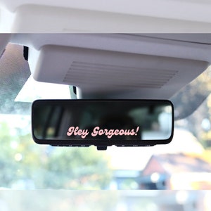 May include: A black rearview mirror with the text "Hey Gorgeous!" in pink script. The mirror is mounted inside a vehicle, with a blurred outdoor scene visible in the reflection.
