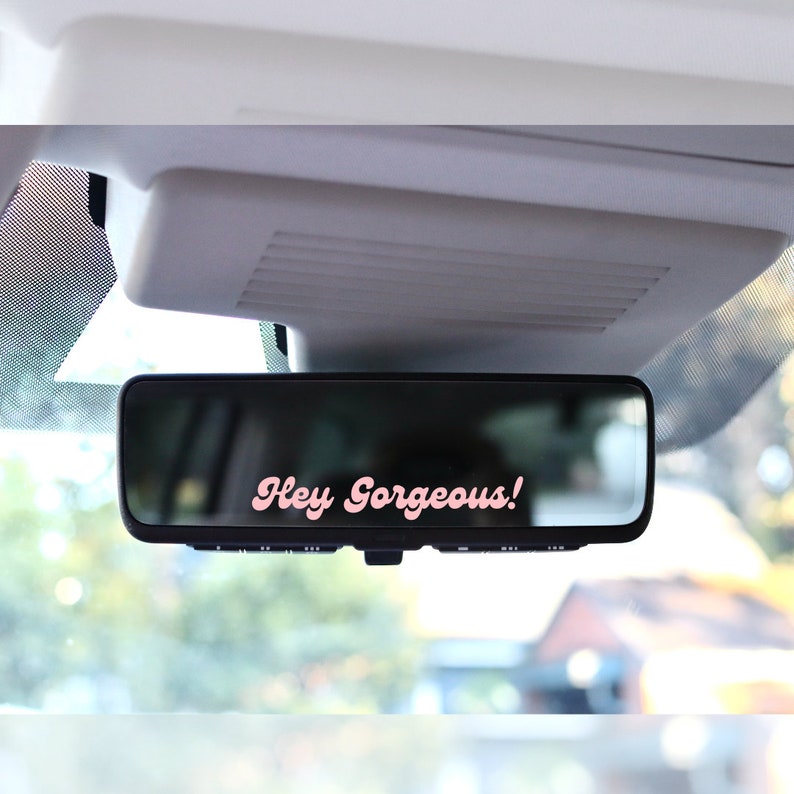 May include: A black rearview mirror with the text "Hey Gorgeous!" in pink script. The mirror is mounted inside a vehicle, with a blurred outdoor scene visible in the reflection.