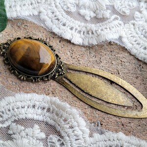 Op de afbeelding: Een gouden bladwijzer met een tijgeroog-steen in een decoratief frame. De bladwijzer is lang ovaalvormig met een puntig uiteinde.