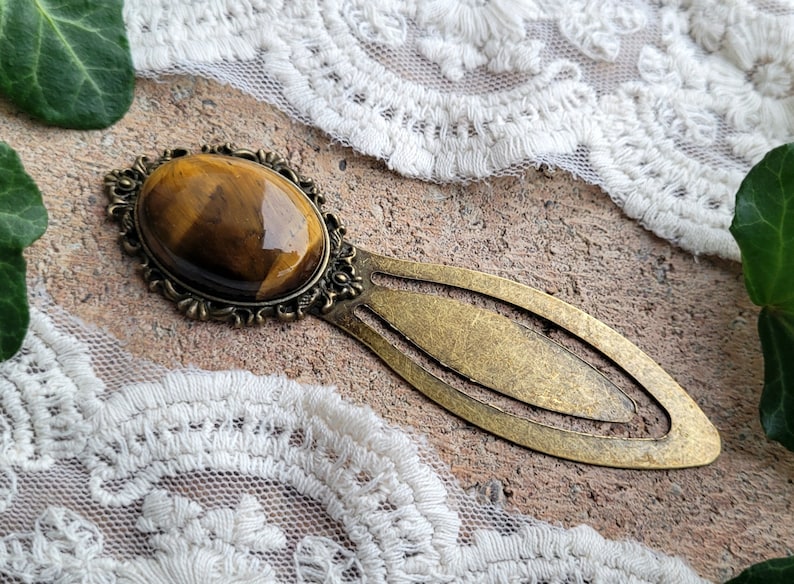 Op de afbeelding: Een gouden bladwijzer met een tijgeroog-steen in een decoratief frame. De bladwijzer is lang ovaalvormig met een puntig uiteinde.