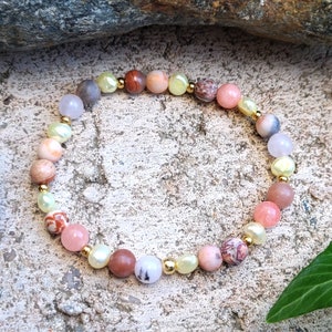 Puede incluir: Un brazalete hecho de piedras de varios colores, incluyendo rosa, marrón y blanco, con detalles dorados. El brazalete está sobre una superficie gris texturizada.
