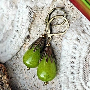 May include: A pair of drop earrings featuring smooth, pear-shaped lime green stones. Each stone is capped with an ornate, antique bronze-toned metal filigree setting and suspended from a matching bronze-toned leverback ear wire closure.