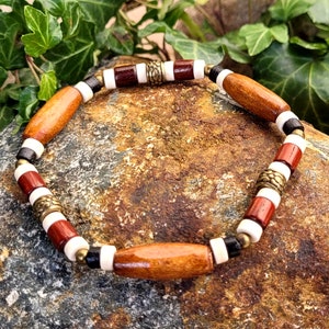 Könnte beinhalten: Ein Holzperlenarmband mit braunen, roten, weißen und schwarzen Perlen. Das Armband hat goldfarbene Metallakzente.