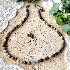 Peut inclure: Un collier de pierres multicolores avec une paire de boucles d'oreilles assorties. Le collier est compos&eacute; de pierres de diff&eacute;rentes nuances de brun, vert et rose. Les boucles d'oreilles sont faites des m&ecirc;mes pierres et ont un design simple.