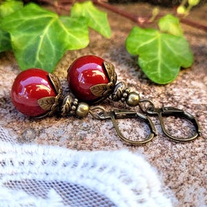 Könnte beinhalten: Ein Paar rote Glasperlenohrringe mit antiken bronzefarbenen Blattakzenten und Bügelverschlüssen.