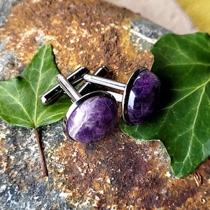 May include: A pair of round cufflinks featuring polished purple amethyst stones set in silver-toned metal frames. The stones display natural veining and a smooth, domed surface, with the metal posts extending from the back of each circular setting.