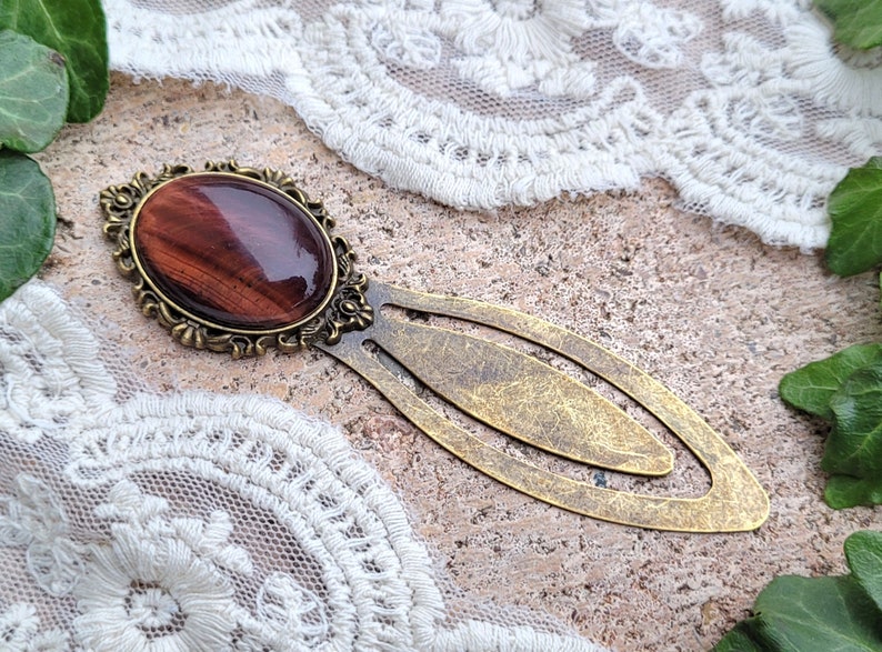 Op de afbeelding: Een gouden bladwijzer met een rode tijgeroog-steen in een decoratief frame. De bladwijzer heeft een ontwerp met dubbele lus.