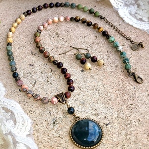 May include: A multi-colored gemstone necklace with a large, round, dark green pendant. The necklace is made with a variety of stones, including tiger's eye, jasper, and agate. The pendant is set in a bronze-colored frame. The necklace is accompanied by a pair of matching earrings.