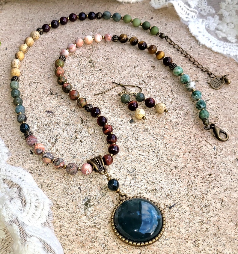 May include: A multi-colored gemstone necklace with a large, round, dark green pendant. The necklace is made with a variety of stones, including tiger's eye, jasper, and agate. The pendant is set in a bronze-colored frame. The necklace is accompanied by a pair of matching earrings.
