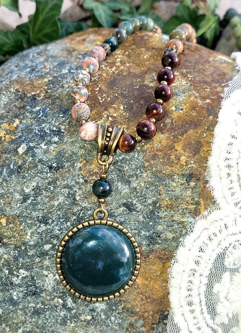 May include: A green agate gemstone pendant necklace with a brown and gold metal setting. The necklace is made with a variety of stones, including red tiger's eye, pink and green jasper, and green moss agate.