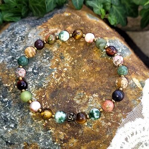 May include: A multi-colored beaded bracelet with a variety of stones, including green, brown, and pink. The bracelet has a gold metal charm in the center.