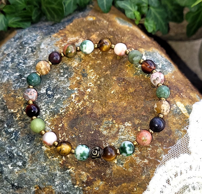 May include: A multi-colored beaded bracelet with a variety of stones, including green, brown, and pink. The bracelet has a gold metal charm in the center.