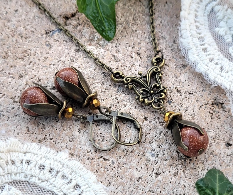 Pode incluir: Um conjunto de brincos e um colar com uma pedra marrom e detalhes dourados. Os brincos t&ecirc;m formato de l&aacute;grima e o colar tem um delicado pingente de borboleta.
