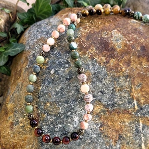 May include: A multi-colored beaded necklace with gold accents. The beads are in shades of pink, green, brown, and red. The necklace is lying on a rock.