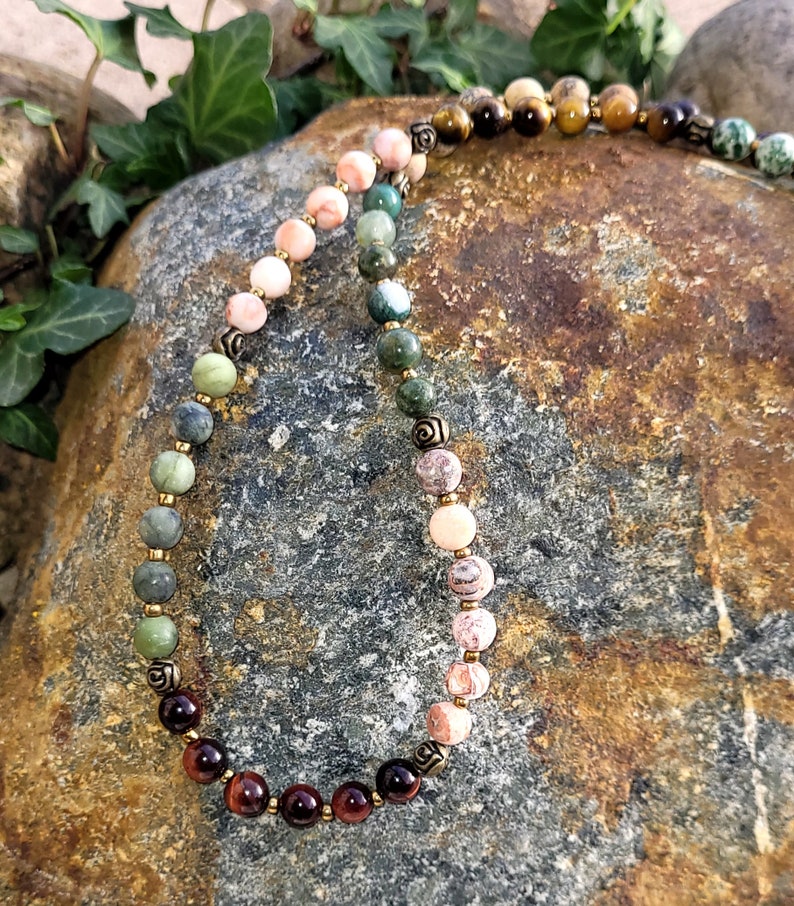 May include: A multi-colored beaded necklace with gold accents. The beads are in shades of pink, green, brown, and red. The necklace is lying on a rock.