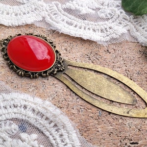 Op de afbeelding: Een gouden bladwijzer met een rode glazen cabochon in een decoratieve setting. De bladwijzer is lang ovaalvormig met een kleinere ovale uitsnijding in het midden.