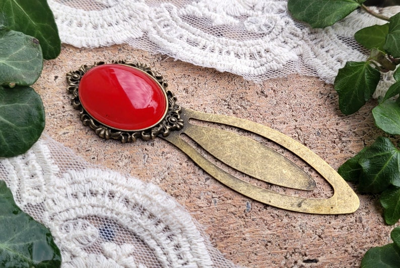 Op de afbeelding: Een gouden bladwijzer met een rode glazen cabochon in een decoratieve setting. De bladwijzer is lang ovaalvormig met een kleinere ovale uitsnijding in het midden.