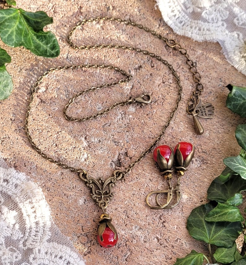 Pode incluir: Um conjunto de colar e brincos de bronze com contas de vidro vermelho em forma de bagas. O colar apresenta um delicado pingente de borboleta e uma corrente com fecho de lagosta. Os brincos s&atilde;o de estilo pendente com um aro pequeno.