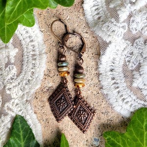 Può includere: Un paio di orecchini color rame con un pendente a forma di diamante. Gli orecchini presentano un motivo decorativo e sono impreziositi da perline colorate. Gli orecchini sono esposti su una superficie strutturata con pizzo e verde sullo sfondo.